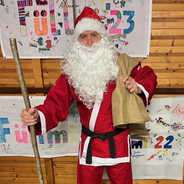 Mann im roten Kostüm mit weißem Bart und Stock, vor kunstvoll beschrifteten Plakaten aus Pappe.