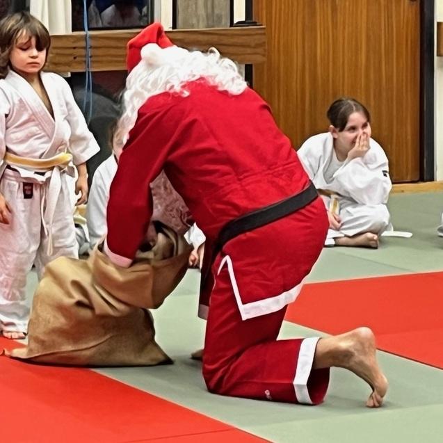 Weihnachtsmann kniet auf dem Boden und übergibt Geschenke an Kinder in Ju-Jutsu-Uniformen in einem Dojo.