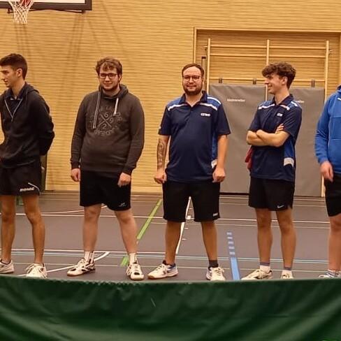 Fünf Männer in Sportkleidung stehen an einem Tischtennistisch in einer Halle. Hintergrund mit Basketballkorb sichtbar.