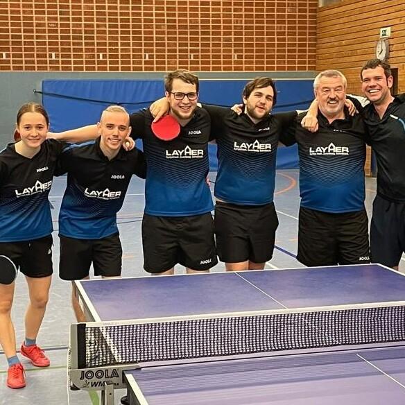 Sechs Tischtennisspieler in einheitlicher Sportkleidung posieren lächelnd vor einem Tischtennistisch in einer Halle.