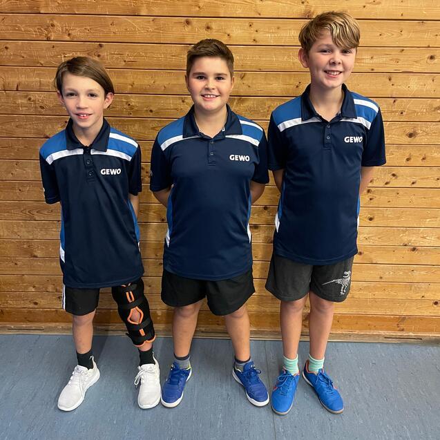 Drei Kinder in blauen Sporttrikots stehen vor einer Holzwand, lächeln und posieren für ein Foto.