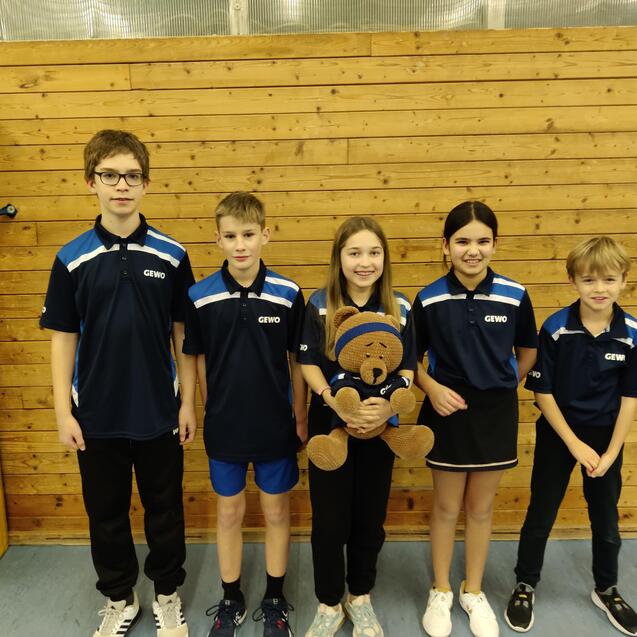 Fünf Kinder in blauen Sporttrikots stehen vor einer Holzwand und halten einen Stoffbären in der Mitte.