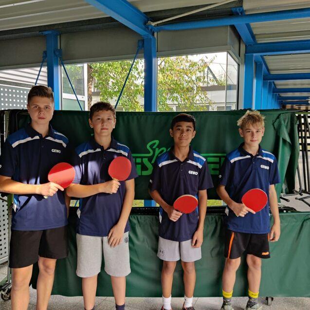 Vier Jungen in blauen T-Shirts stehen vor einem Tischtennistisch und halten Tischtennisschläger mit roten Belägen.