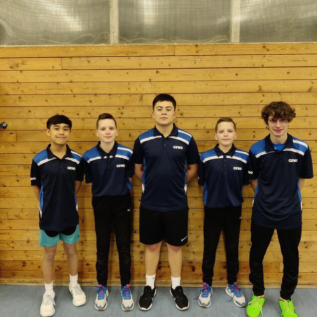 Fünf Kinder in blauen Tischtennisshirts stehen vor einer Holzwand in einer Sporthalle.