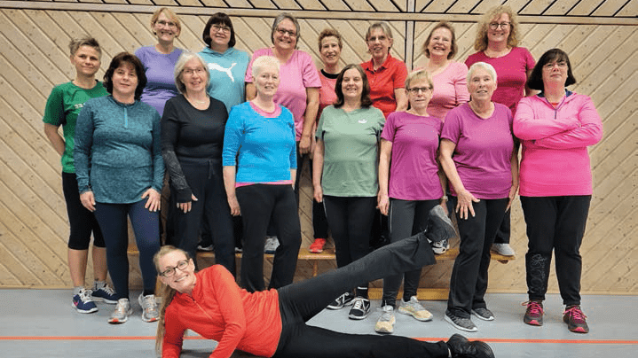 Gruppenzusammenstellung von 18 Frauen in Sportkleidung, die l&auml;chelnd in einer Turnhalle posieren. Eine Frau liegt im Vordergrund.