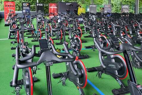 Reihe von stationären Fahrrädern in einem überdachten Bereich mit grüner Bodenbelag und Werbebannern im Hintergrund.