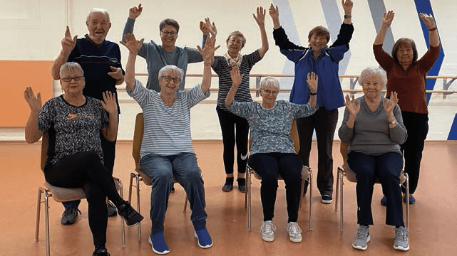 Eine Gruppe von zehn Senioren sitzt und steht, hebt fr&ouml;hlich die H&auml;nde in die Luft, in einem &Uuml;bungsraum.