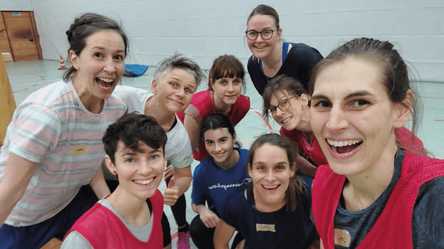 Gruppe von neun Frauen in Sportkleidung l&auml;chelt und posiert gemeinsam in einer Sporthalle.