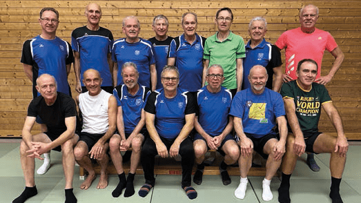 Gruppe von M&auml;nnern in Sportkleidung, die auf einer Matte sitzen, l&auml;cheln und f&uuml;r ein Foto posieren.