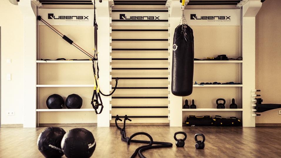 Fitnessraum mit einem Sandsack, Kettlebells, Matzentrainern und Trainingsgeräten auf Regalen vor Holzfußboden.