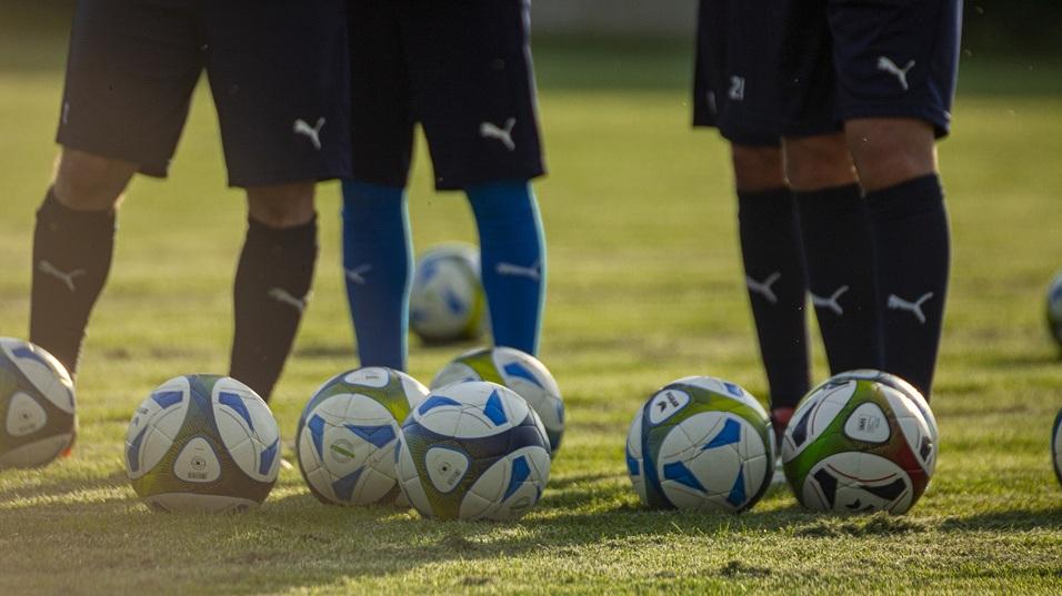Nahaufnahme mehrerer Fußbälle auf einem Rasenplatz vor den Beinen von spielenden Fußballspielern.