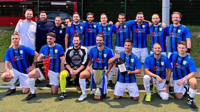 Gruppe von 15 Fußballspielern in blauen Trikots, posierend mit Pokal und Preisen auf einem Sportplatz.