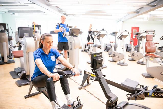Frau im blauen Shirt trainiert auf einem Rudergerät in einem modernen Fitnessstudio mit mehreren Trainingsgeräten im Hintergrund.