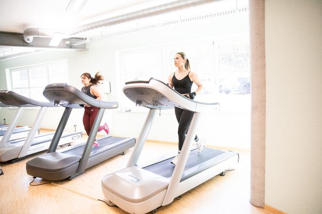 Zwei Frauen joggen auf Laufbändern in einem hellen Fitnessstudio mit großen Fenstern.