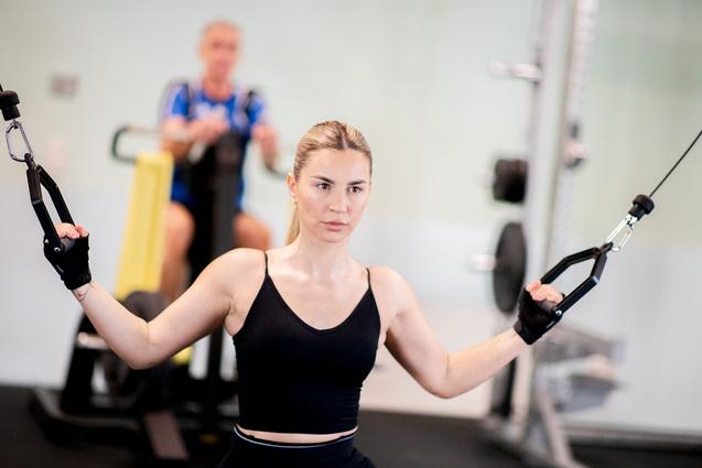 Junge Frau mit Sporthandschuhen trainiert an einem Kabelzuggerät im Fitnessstudio, im Hintergrund eine andere Person.