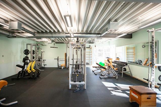 Fitnessstudio mit verschiedenen Geräten, Hanteln und einer großen Fensterfront für Tageslicht.