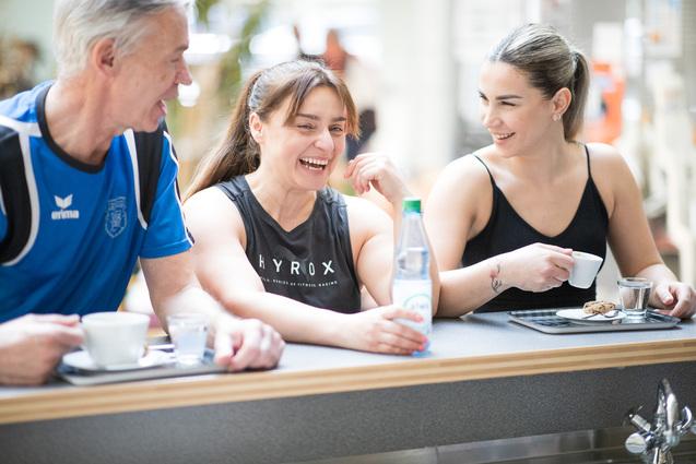 Drei Personen lachen und unterhalten sich an einem Tisch mit Getränken und Snacks in einem freundlichen Ambiente.