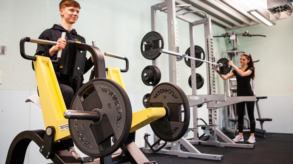 Junger Mann trainiert an einer Kraftmaschine, während eine Frau mit einer Langhantel im Hintergrund trainiert.
