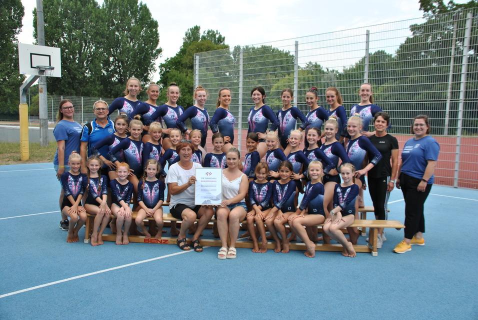 Gruppenfoto von Turnerinnen in blauen Trikots, die stolz ein Zertifikat halten, zusammen mit Trainerinnen auf einem Sportplatz.