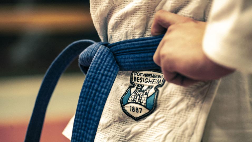 Nahaufnahme einer Hand, die einen blauen Gürtel um einen weißen Judoanzug mit Logo der Sportvereinigung Besigheim bindet.