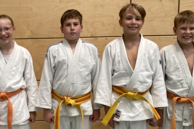 Vier Kinder in weißen Judo-Anzügen mit orangen und gelben Gürteln stehen lächelnd vor einer Holzwand.