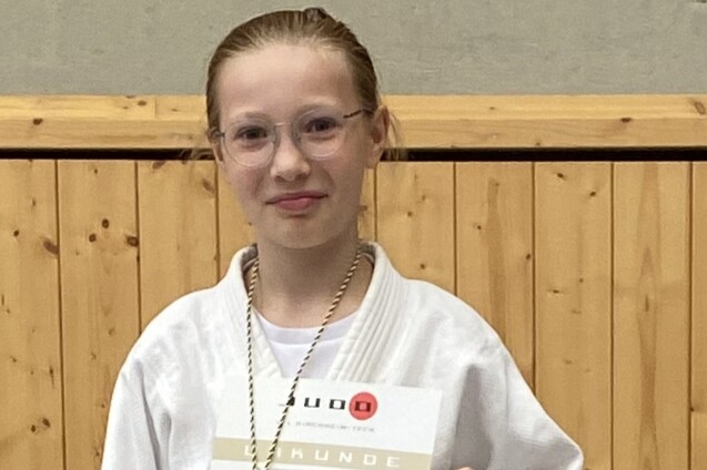 Mädchen in Judoweste hält Urkunde in einer Sporthalle vor Holzwand. Transparentes Schutzschild und Brille sind sichtbar.