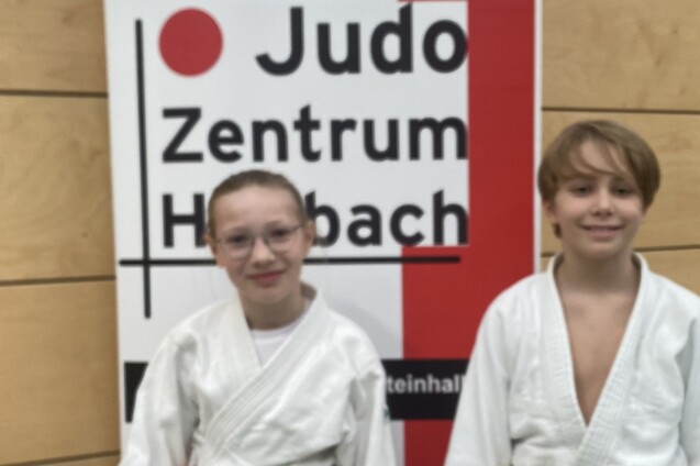 Zwei Kinder in Judoanzügen stehen vor einem Banner des Judozentrums Hohbach, lächeln in die Kamera.