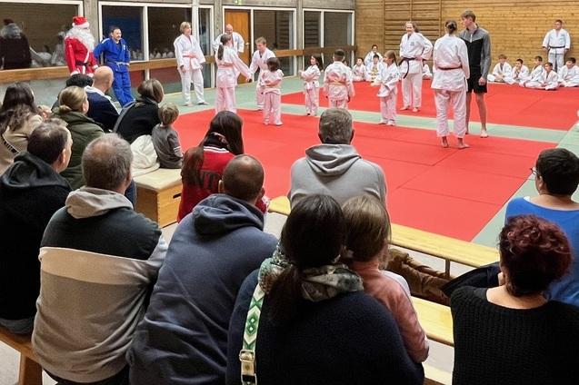 Zuschauer beobachten eine Judo-Vorführung, während Teilnehmer in weißen und blauen Gis auf einer roten Matte trainieren.