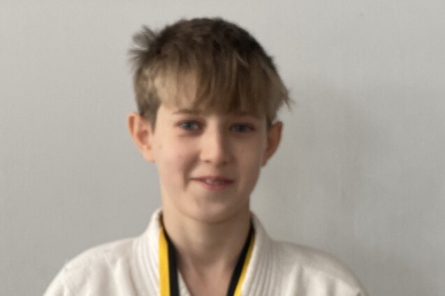 Junge mit blonden Haaren trägt einen weißen Judogi und eine Medaille um den Hals, steht vor einer grauen Wand.