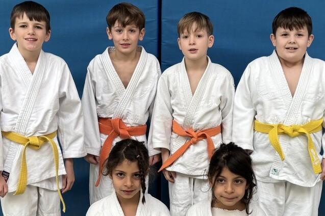 Sechs Kinder in Judokleidung stehen in zwei Reihen vor einer blauen Wand. Zwei tragen orange, zwei gelbe Gürtel.