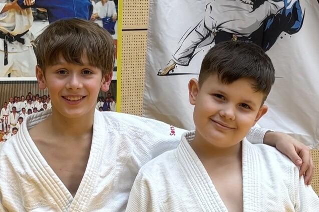 Zwei Kinder in Judo-Gis stehen lächelnd vor einer Wand mit Judo-Fotos und einem Judo-Poster.
