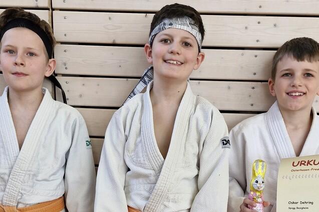 Drei Kinder in Judogis stehen lächelnd vor einer Holzwand, eines hält eine Urkunde und eine Schokoladenfigur.