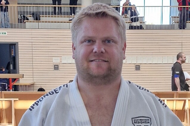 Nahaufnahme eines Mannes in einem wei&szlig;en Judo-Gi, im Hintergrund sind Zuschauer in einer Halle sichtbar.