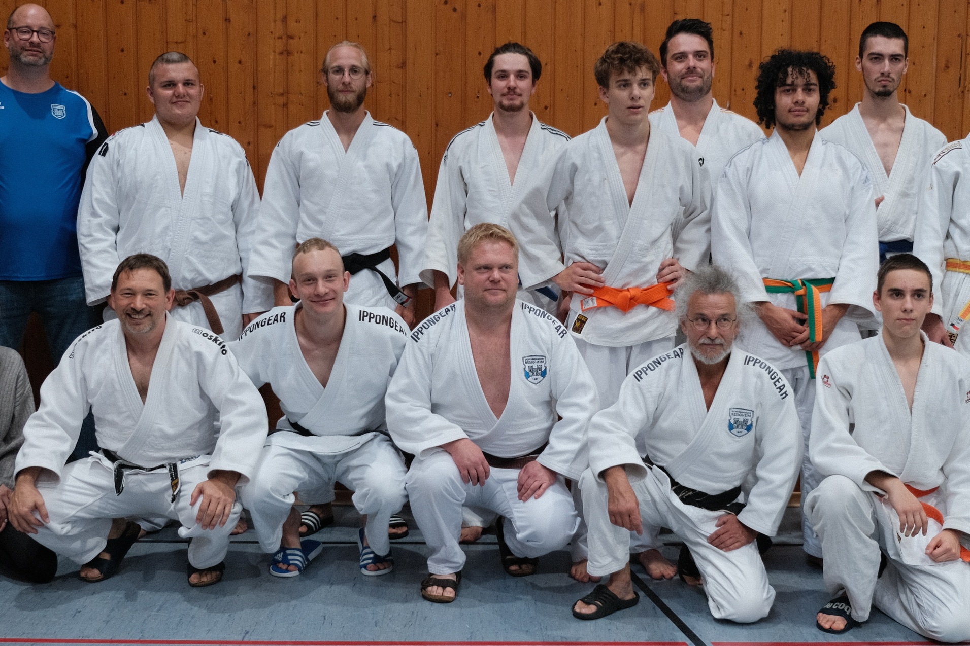 Gruppenfoto von 14 Judoka in weißem Gi, einige mit farbigen Gurten, vor einer Holzpaneel-Wand in einer Sporthalle.