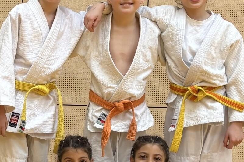 Fünf Kinder in Judo-Anzügen posieren lächelnd vor einer Wand, zwei tragen orange, einer gelbe, zwei weiße Gürtel.
