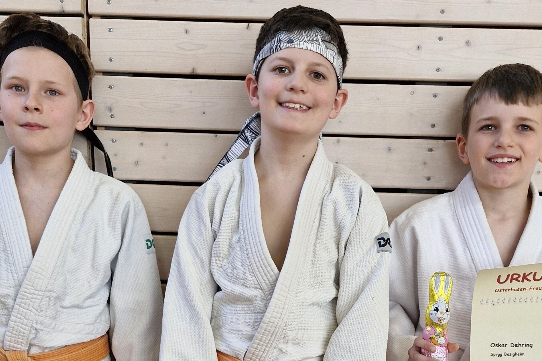 Drei Kinder in Judogis stehen lächelnd vor einer Holzwand, eines hält eine Urkunde und eine Schokoladenfigur.