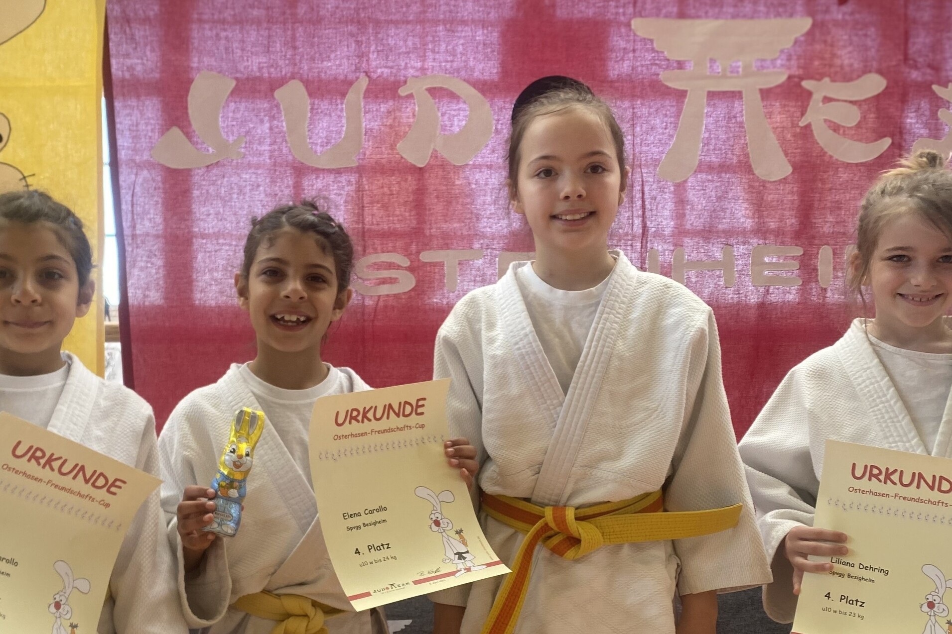 Drei Kinder im Judogi halten Urkunden und Schokoladenhasen, vor einer rot-gelben Wand mit einem Hasenmotiv.