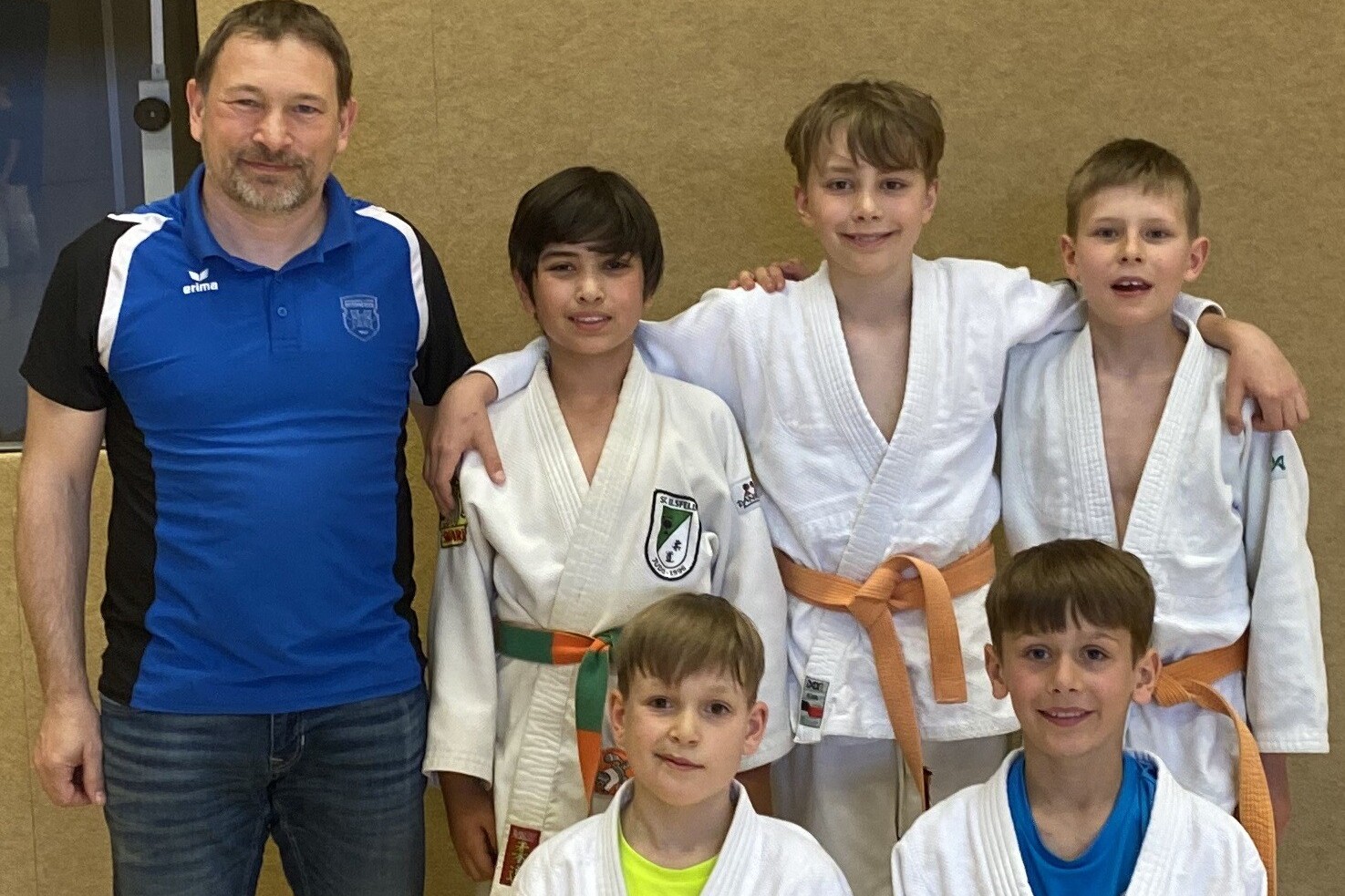 Eine Gruppe von fünf Jungen in Judokleidung steht zusammen mit einem Trainer im Hintergrund, alles vor einer Holzwand.