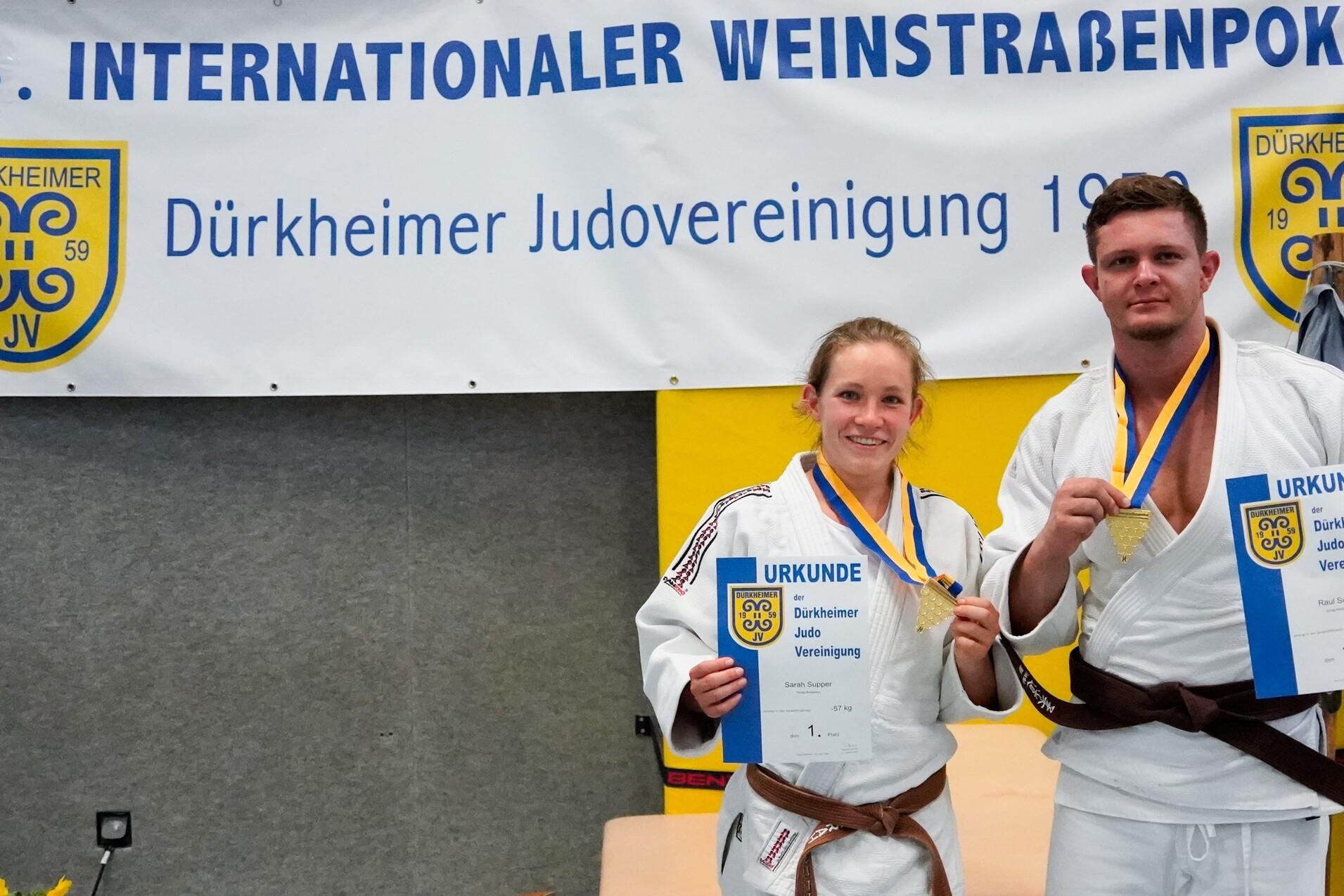 Zwei Judoka posieren stolz mit Medaillen und Urkunden vor einem Banner des Internationalen Weinstraßenpokals.