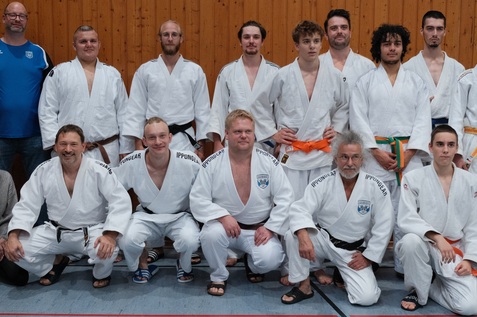 Gruppenfoto von 14 Judoka in wei&szlig;em Gi, einige mit farbigen Gurten, vor einer Holzpaneel-Wand in einer Sporthalle.