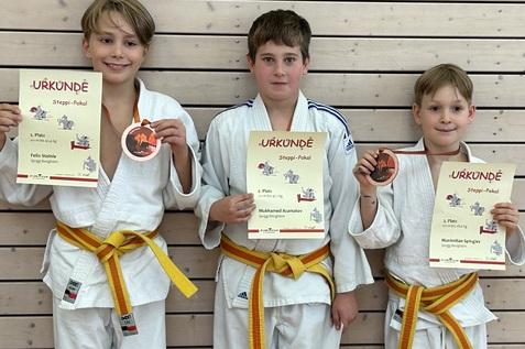 Drei Jugendliche in Judokleidung halten Urkunden und Medaillen des Steppi-Pokals vor einer Holzrückwand.