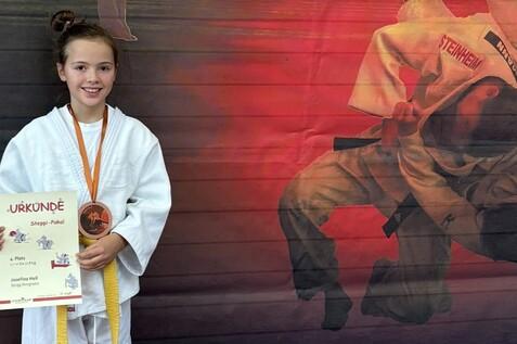 Mädchen in weißem Judogi hält Urkunde und Medaille vor einem Hintergrund mit Judoka in Aktion.