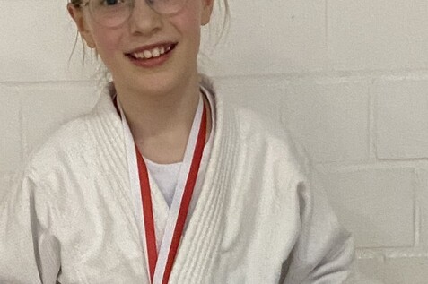 M&auml;dchen in Judokleidung h&auml;lt Urkunde und Medaille vom Kappelbergturnier 2024, l&auml;chelt vor einer wei&szlig;en Wand.