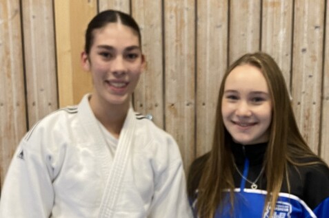 Zwei Jugendliche stehen vor einer Holzwand, eine tr&auml;gt einen wei&szlig;en Judoanzug, die andere eine Sportjacke.