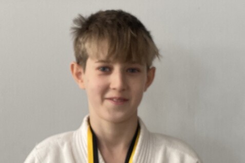 Junge mit blonden Haaren trägt einen weißen Judogi und eine Medaille um den Hals, steht vor einer grauen Wand.