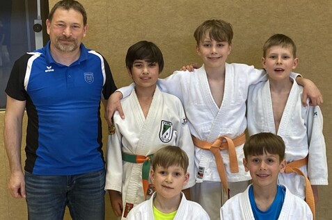 Eine Gruppe von fünf Jungen in Judokleidung steht zusammen mit einem Trainer im Hintergrund, alles vor einer Holzwand.
