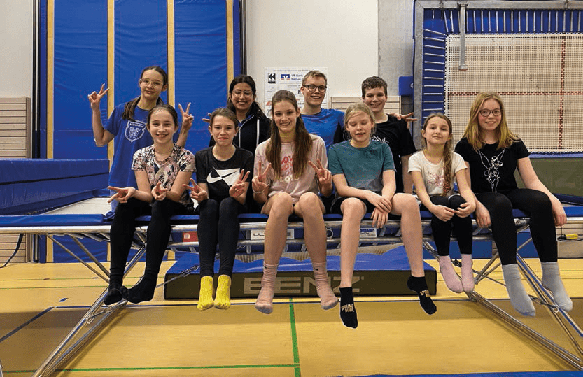 Gruppe von zehn Kindern und Jugendlichen sitzt auf einem Trampolin, lächelnd und mit V-Zeichen.