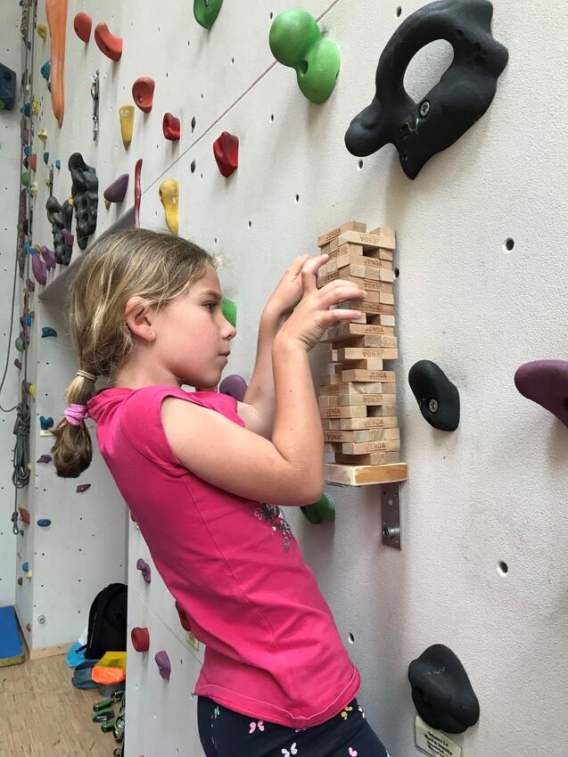 Mädchen in pinkem Shirt baut Jenga-Turm an einer Kletterwand mit bunten Griffen.