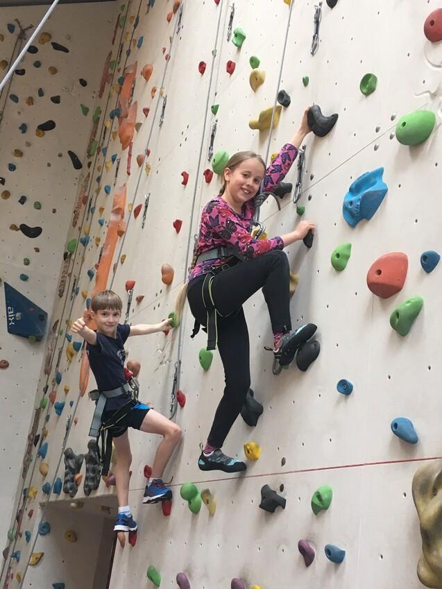 Zwei Kinder klettern an einer Kletterwand mit bunten Griffen, lächelnd und in Sicherungsgurten.