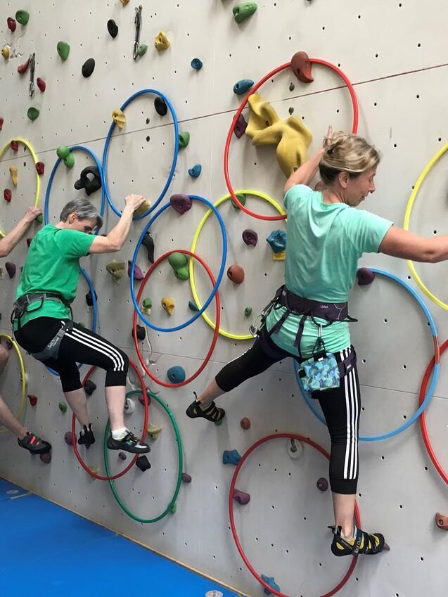 Drei Personen klettern an einer bunten Kletterwand mit Ringen und verschiedenen Griffen.